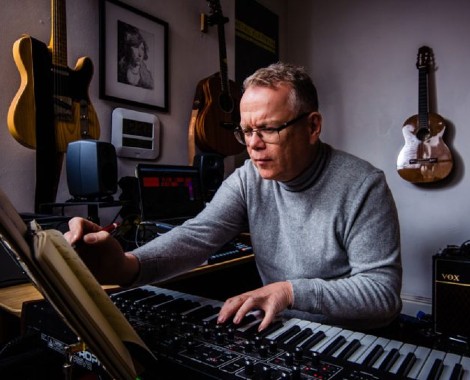 A Caucasian male wearing glasses and a grey jumper plays piano in a room with guitars hanging on the wall.